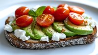 Fresh avocado toast topped with sliced cherry tomatoes, crumbled feta cheese, basil leaves and black pepper on rustic sourdough bread, served on white plate.