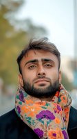 Middle Eastern man with beard wearing colorful floral scarf and black jacket outdoors, looking confidently at camera against blurred background. Lifestyle portrait.