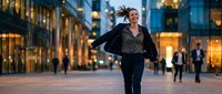 Cheerful young Caucasian businesswoman running joyfully on urban city street at dusk, jacket in hand, modern glass buildings in background.