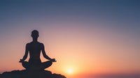 Silhouette of woman meditating in lotus pose against stunning sunset sky with gradient colors from blue to orange for wellness and mindfulness concepts.