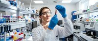 Female scientist in white lab coat and blue gloves examining test tube in modern laboratory with equipment and supplies. For research, healthcare, and scientific publications.