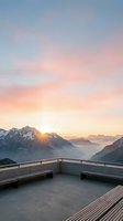 Sunrise over snow-capped mountains viewed from modern concrete terrace with metal railing, misty valley below, golden light breaking through clouds, ideal for travel and lifestyle design projects.