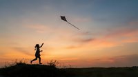 Child flying kite at sunset on hilltop, silhouetted against golden sky. Outdoor recreation and leisure activity for kids.