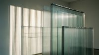 Young adult woman with light skin tone and blonde hair, wearing casual clothing, standing in a modern office or residential space with floor-to-ceiling glass partitions and vertical blinds, natural daylight streaming through windows creating geometric shadows.