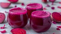 Fresh beetroot smoothie in glass cups surrounded by raw sliced beets and scattered petals on light surface. Vibrant magenta drink for healthy lifestyle.