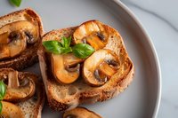 Grilled mushroom bruschetta with fresh basil on toasted bread slices, served on gray ceramic plate against white marble background. Appetizing Italian starter.