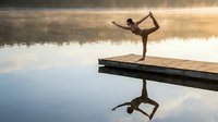 Young woman practicing yoga dancer pose on wooden dock at serene lake during golden hour sunrise with perfect water reflection creating peaceful meditation scene.
