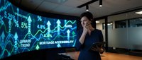 Young woman with dark hair and medium skin tone, wearing professional navy blazer, standing in modern office environment while analyzing financial data on large curved digital display showing upward trends, percentages, and mortgage accessibility charts, holding tablet device.