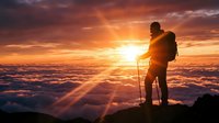 Silhouette of male hiker with backpack and trekking poles standing on mountain peak at golden sunrise, watching sun rays breaking through clouds below.