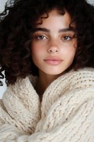 Young woman with natural curly dark hair and freckles wearing cozy cream knit sweater, looking directly at camera with soft natural makeup and gentle expression.
