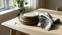 Stack of ceramic plates with flour dusting and linen cloth on wooden table near window, ready for baking or cooking preparation in bright kitchen setting.