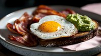 Sunny side up egg on dark toast with crispy bacon strips and fresh avocado spread served on gray ceramic plate, close up view of breakfast meal.