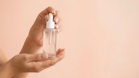 Hands holding clear glass dropper bottle with white pipette cap against warm beige background for skincare, cosmetics, serum, and beauty product packaging.
