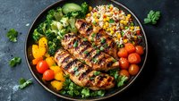 Grilled chicken breast with colorful rice, cherry tomatoes, corn, broccoli and fresh herbs served on dark plate against textured background for restaurant menu.