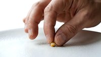 Adult male hand with medium skin tone picking up a small gold nugget or precious metal fragment from white surface, representing wealth, mining, investment, and precious metals commerce.