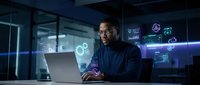 African American male cybersecurity analyst working on laptop with holographic data visualizations in modern tech office environment at night.