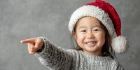 Cheerful Asian girl in Santa hat pointing with excitement, wearing cozy gray sweater against neutral background. Holiday portrait captures childhood joy.