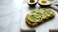 Healthy avocado toast on rustic bread with seeds and herbs on marble surface. Fresh breakfast meal with sliced avocados in background for nutritious eating.