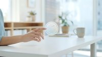 Woman's hand holding a white jar of skincare cream on a bright, minimalist desk with potted plants and windows in background, suitable for beauty, cosmetics, and wellness marketing.
