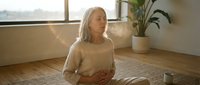 Mature Caucasian woman practicing mindful meditation in bright modern living room with natural sunlight streaming through windows.