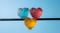 Colorful heart-shaped ice cubes in blue, red and yellow hanging on black rod against light blue background, frozen water drops creating romantic mood.