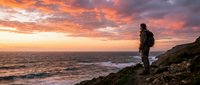 Male hiker with backpack standing on rocky coastal cliff at sunset, watching ocean waves and dramatic sky. Adventure travel and outdoor exploration concept.