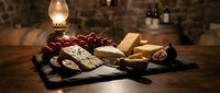 Assorted cheeses and red grapes arranged on slate board with vintage oil lamp and wine bottles in rustic cellar setting for food and wine pairing.