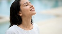Young Hispanic woman with wet hair smiling peacefully with eyes closed outdoors near water, expressing joy and relaxation in natural summer setting.