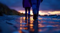 Silhouetted figures walking on wet beach at sunset, with dramatic reflections of orange sky in water puddles creating magical atmosphere and bokeh effect.