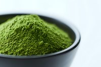 Vibrant green matcha tea powder in black ceramic bowl against light background, selective focus on fine powder texture showing authentic Japanese tea quality.