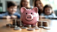 Pink ceramic piggy bank with happy smiling face surrounded by stacks of silver coins on wooden table, representing savings, financial education and money management.