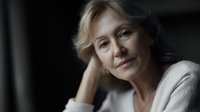 Mature blonde woman resting chin on hand with gentle smile and contemplative expression in soft natural lighting for lifestyle and wellness content.