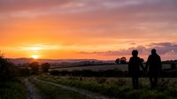 Two people standing on a rural path watching golden sunset over countryside landscape with mountains in distance, suitable for lifestyle and nature editorial use.