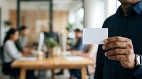 Man in dark navy shirt holding blank white business card in modern office environment with colleagues working at wooden table in background.