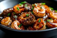 Glazed shrimp with sesame seeds and green onions served in black plate. Close up view of asian style seafood with sweet and savory sauce garnished with herbs.