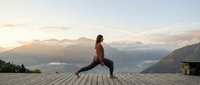Woman in brown jacket performing yoga lunge on wooden deck overlooking misty mountain valley at sunrise, wellness and outdoor fitness concept.