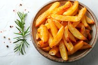 Crispy baked potato wedges seasoned with herbs and spices in rustic ceramic bowl, fresh rosemary sprig and red pepper flakes on light background.
