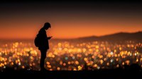 Silhouette of person using smartphone against orange sunset sky with blurred city lights bokeh background. Urban evening technology concept.