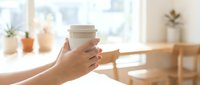 Hands holding takeaway coffee cup in bright cafe interior with natural sunlight streaming through windows creating warm atmosphere for morning beverage enjoyment.