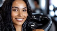 Young African American female hairdresser with bright smile and long dark hair wearing black gloves in modern salon interior with blurred background. Professional beauty service.