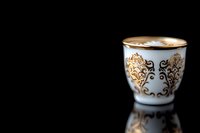 Elegant white porcelain coffee cup with ornate gold floral pattern and fresh cappuccino foam art on dark reflective surface against black background.