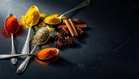Colorful spices and seasonings arranged on dark slate background with vintage spoons. Culinary ingredients for cooking and food preparation.