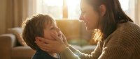 Loving mother gently holding her young son's face in warm sunlit living room, showing tender family bonding moment and maternal care at home.