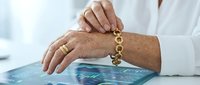 Senior woman with light skin tone wearing gold jewelry, holding a dangling gold chain bracelet over a digital tablet displaying financial data, representing wealth management and investment planning.