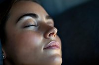 Young Asian woman with closed eyes in peaceful meditation pose, face illuminated by soft side lighting against dark background, showing serenity and mindfulness.