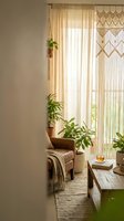 Cozy boho living room corner with macrame wall hanging, sheer curtains, houseplants and natural textures creating warm minimalist home decor atmosphere.