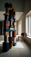 Modern architectural wooden tower sculpture with stacked geometric blocks in blue, red, yellow, and natural wood tones, displayed in minimalist interior space with mid-century chair and bright windows.