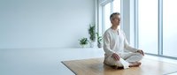 Young Caucasian woman in white clothing practicing meditation in modern minimalist room with large windows and natural light for wellness and mindfulness.