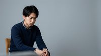 Young Asian man in dark navy sweater sitting on wooden chair against neutral gray background, looking down with contemplative expression, suitable for business, lifestyle, and editorial use.