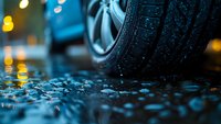 Car tire on wet asphalt during rain with bokeh city lights in background creating moody atmospheric night scene. Closeup view shows water droplets and reflections.